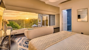 A bedroom with a large bed, a seating area with a chair and a table, a view of the outdoors, and a door leading to another room.