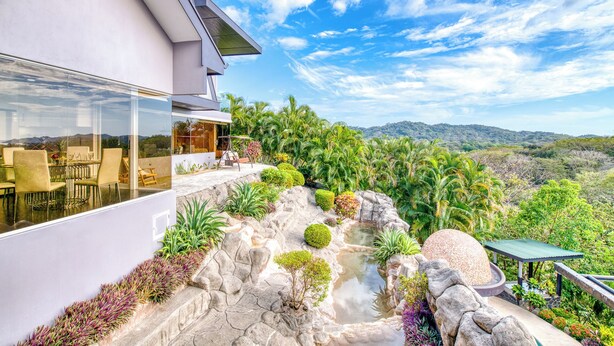 A modern house with a dining area overlooking a lush garden and a scenic mountain view.