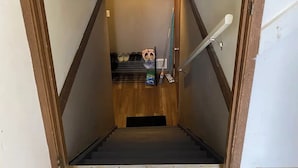 Un escalier étroit avec une main courante en bois, une poignée de porte en métal et une petite étagère où sont rangées des chaussures et des produits de nettoyage.