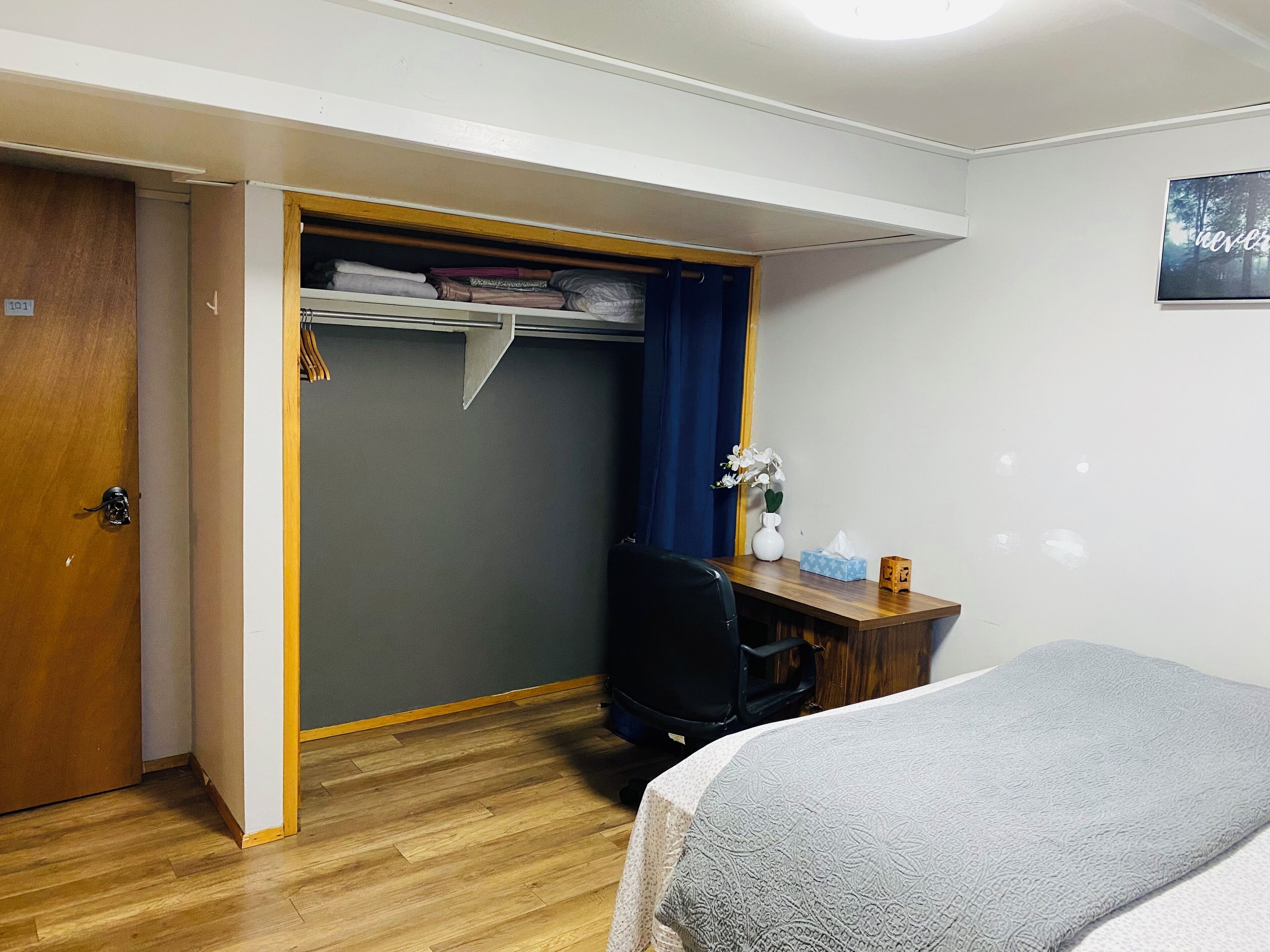 A hotel room with a bed, a desk with a chair, a wardrobe, and a small table with a vase and tissues.