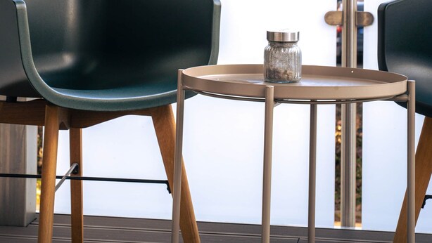 Un balcon avec deux chaises noires et une petite table, d’où l’on peut admirer un paysage urbain avec des immeubles et le clocher d’une église.