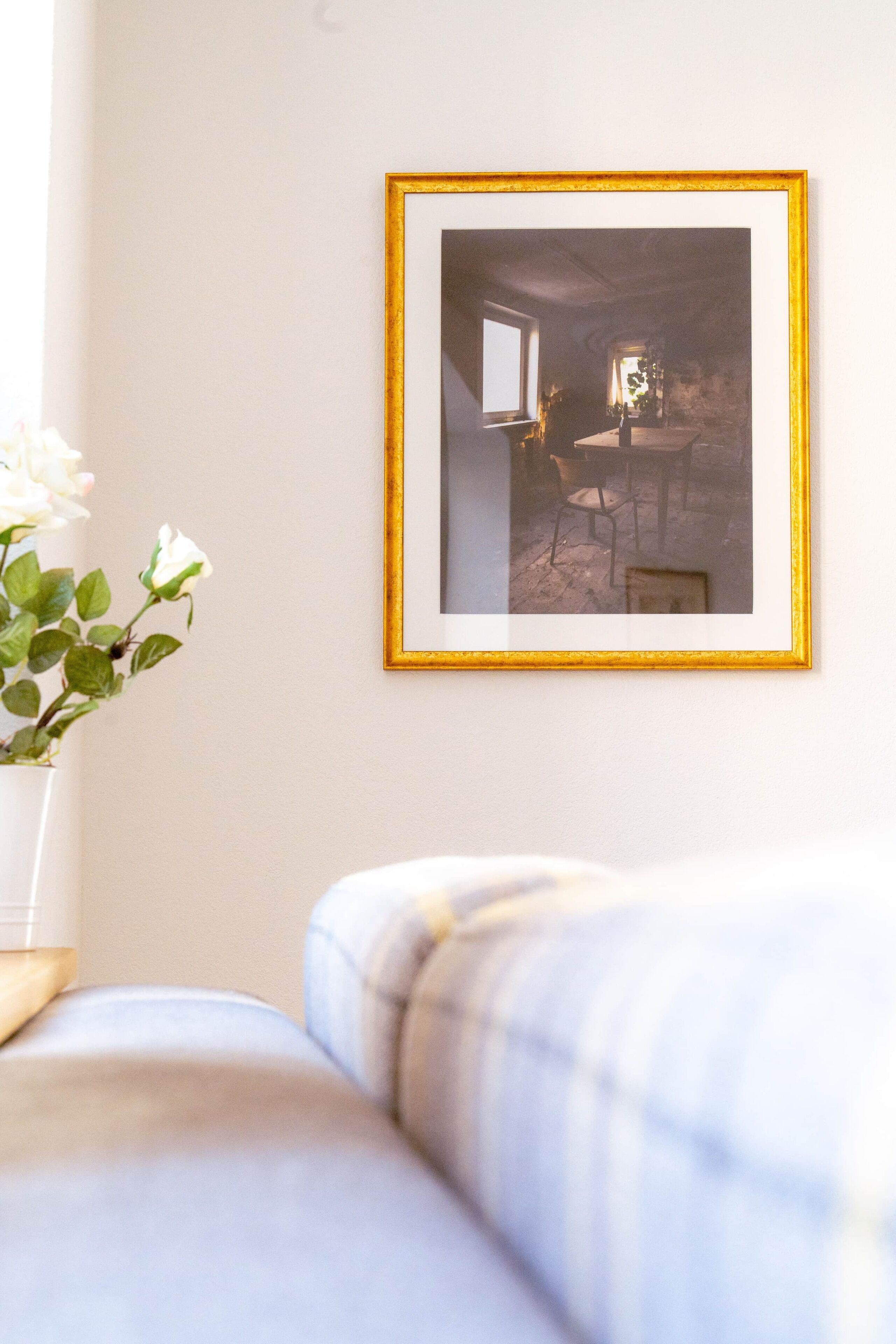 A framed photograph on a wall, a white vase with white flowers, and a light blue sofa.
