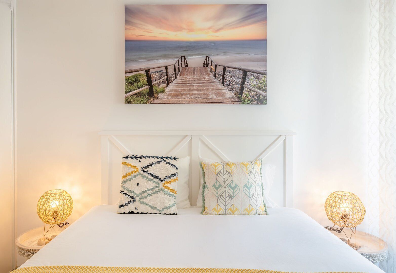 Une chambre avec un lit en bois, deux coussins à motifs, un tableau représentant un coucher de soleil sur la plage au-dessus du lit, et deux lampes de chevet.