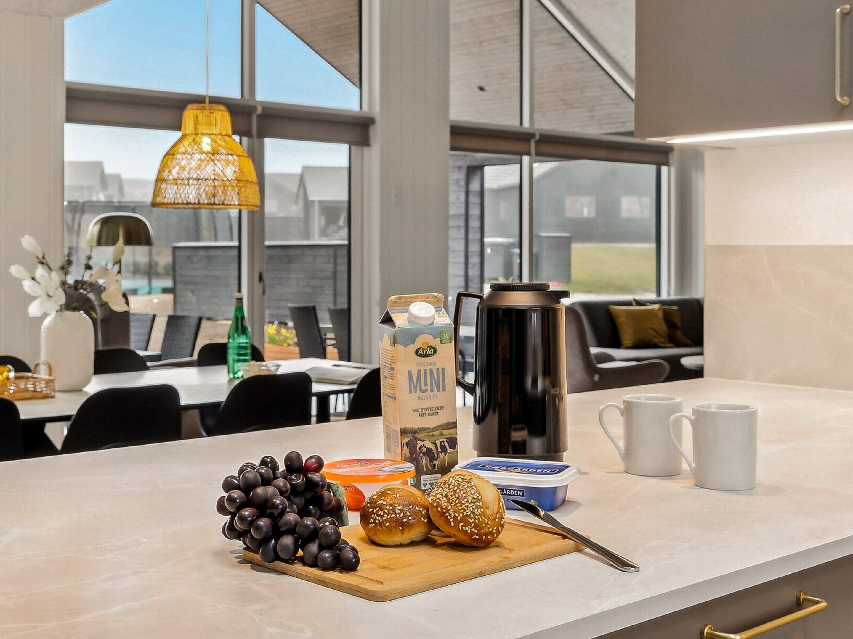 Eine moderne Küche mit Frühstücksecke, darunter eine Milchpackung, eine Kaffeemaschine, ein Schneidebrett mit Brot und Trauben sowie ein Essbereich im Hintergrund.