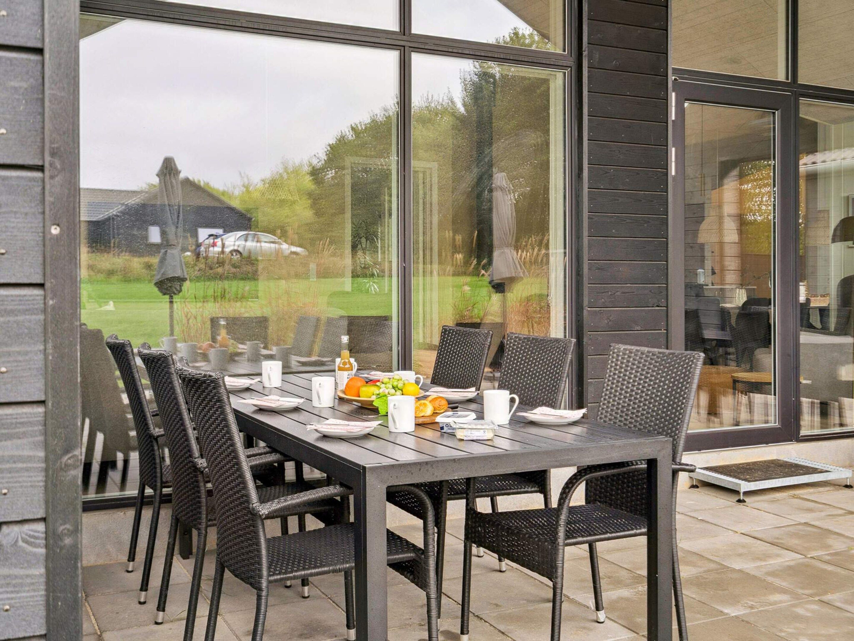 Un espace de restauration en plein air avec une table dressée pour quatre personnes, entourée de chaises en osier, et de grandes fenêtres offrant une vue sur l’extérieur.