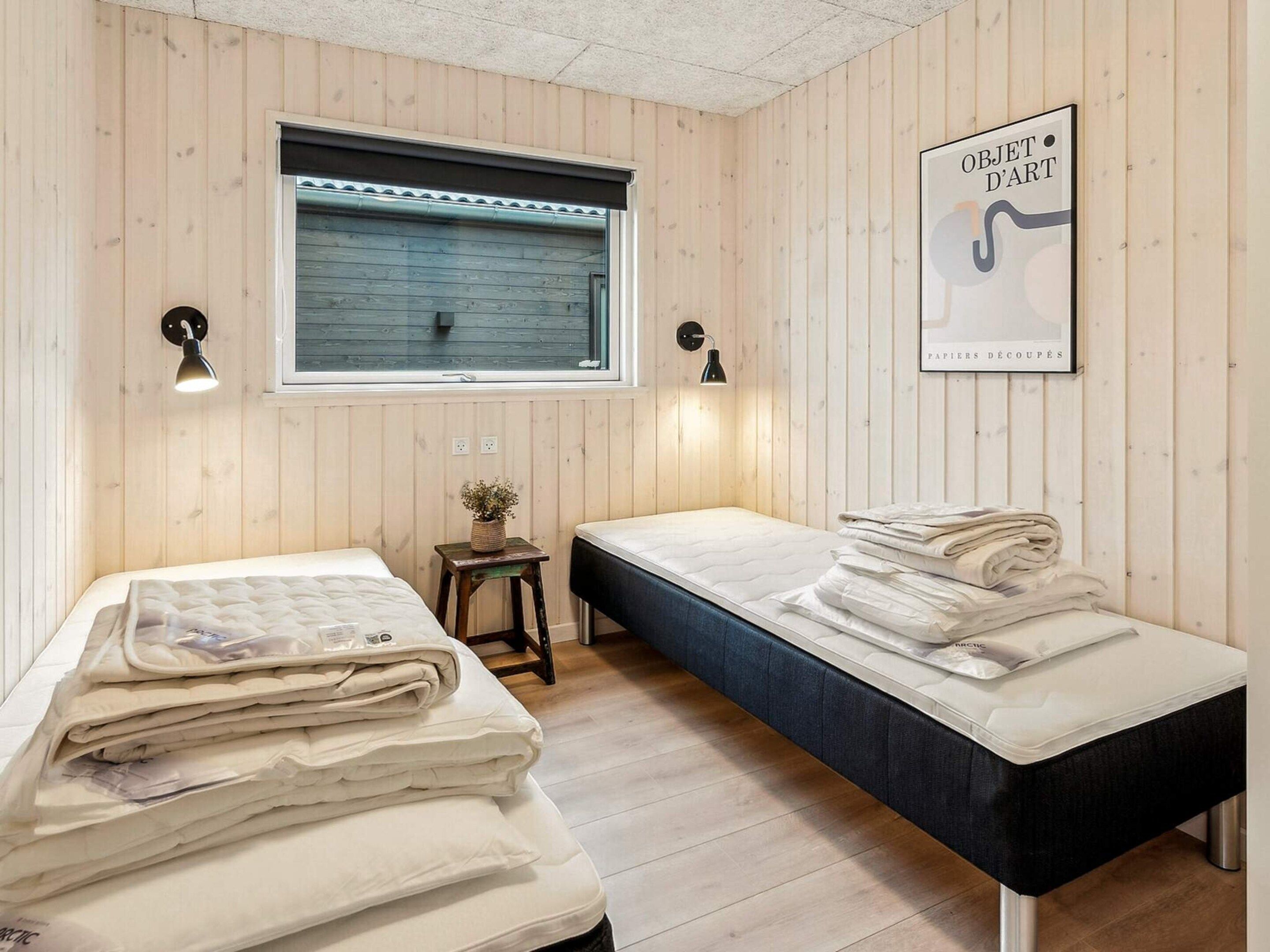 A room with two beds, a window with blinds, a small table with a plant, and wall-mounted lamps.