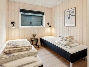 A room with two beds, a window with blinds, a small table with a plant, and wall-mounted lamps.