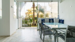 Een moderne eethoek met een glazen tafel en metalen stoelen, een balkon met een glazen deur en uitzicht op palmbomen en gebouwen.