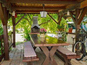 Una pérgola de madera al aire libre con un horno de pizza de piedra, una mesa de picnic, bancos y plantas en macetas.