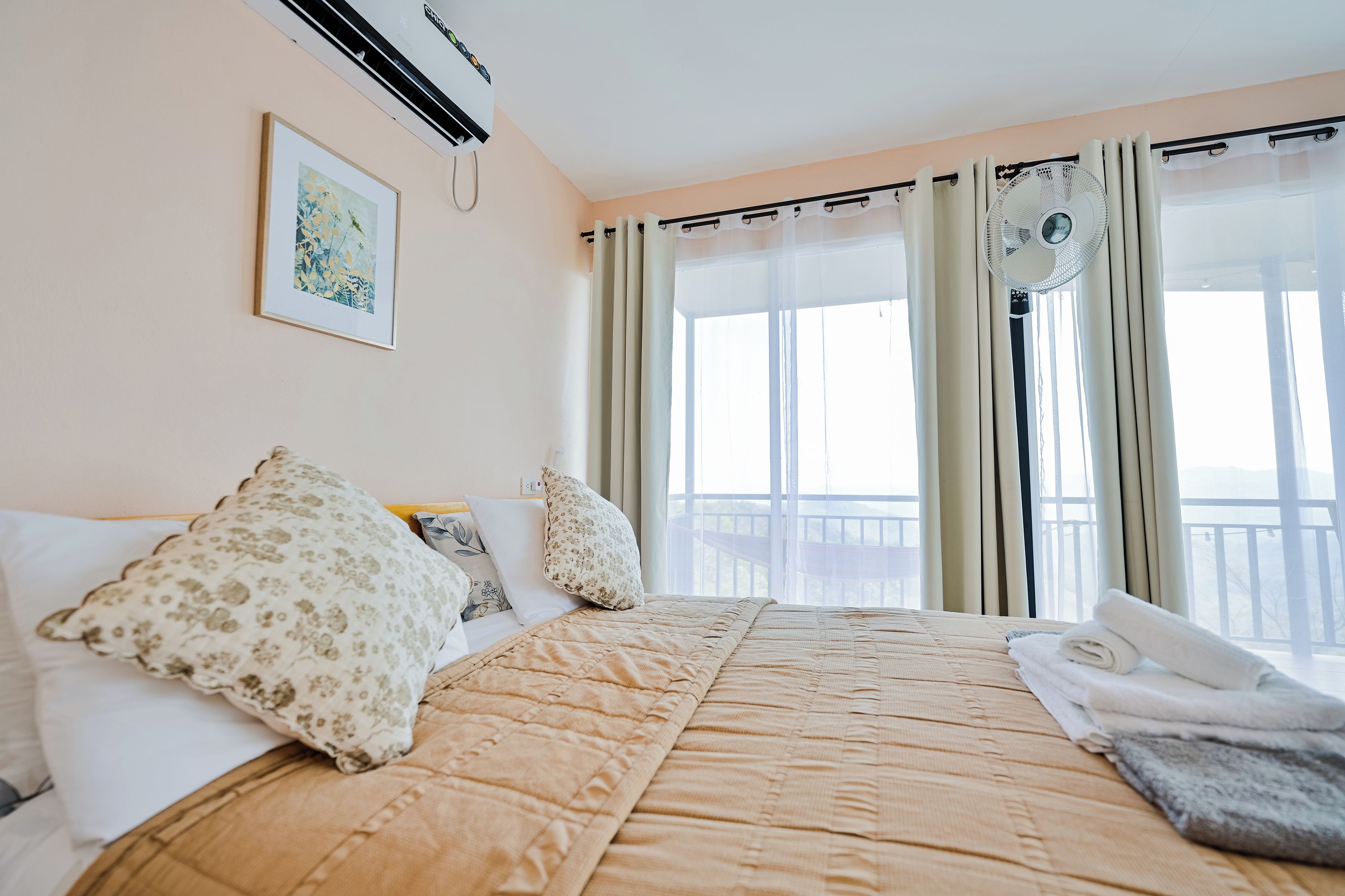 A hotel room with a bed, a window with curtains, a wall-mounted air conditioner, and a framed picture.