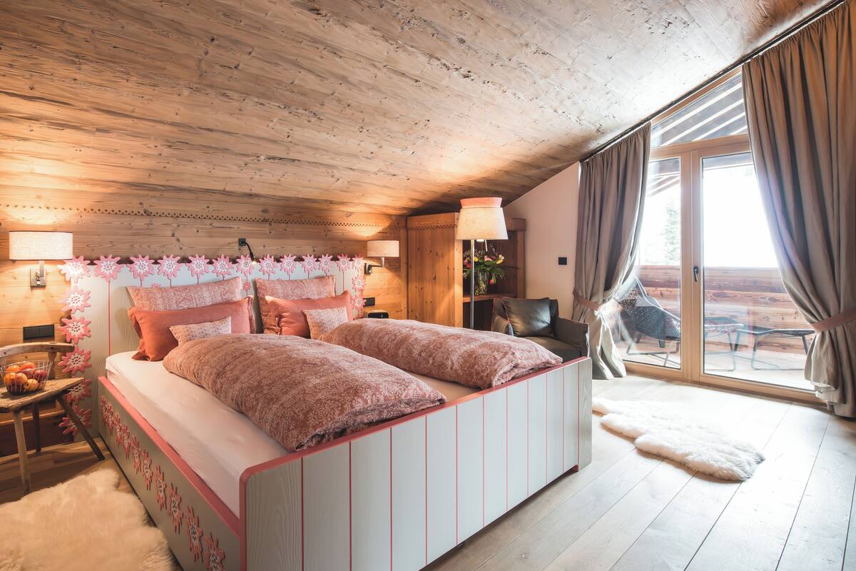 Ett sovrum med ett träpanelat tak, en stor säng med rosa sängkläder, ett nattduksbord med en lampa och utsikt över naturen genom en skjutdörr i glas.