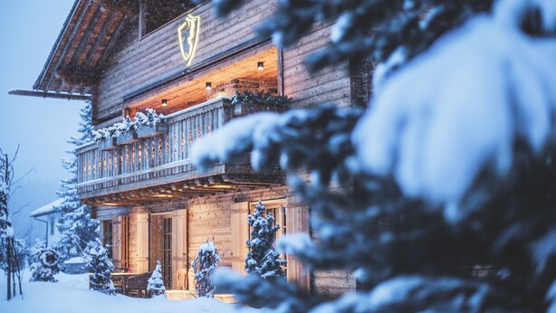 A wooden chalet with a balcony, snow-covered trees, and a lit sign above the entrance. - Lech Lodge - LEGENDARY (Lech am Arlberg)