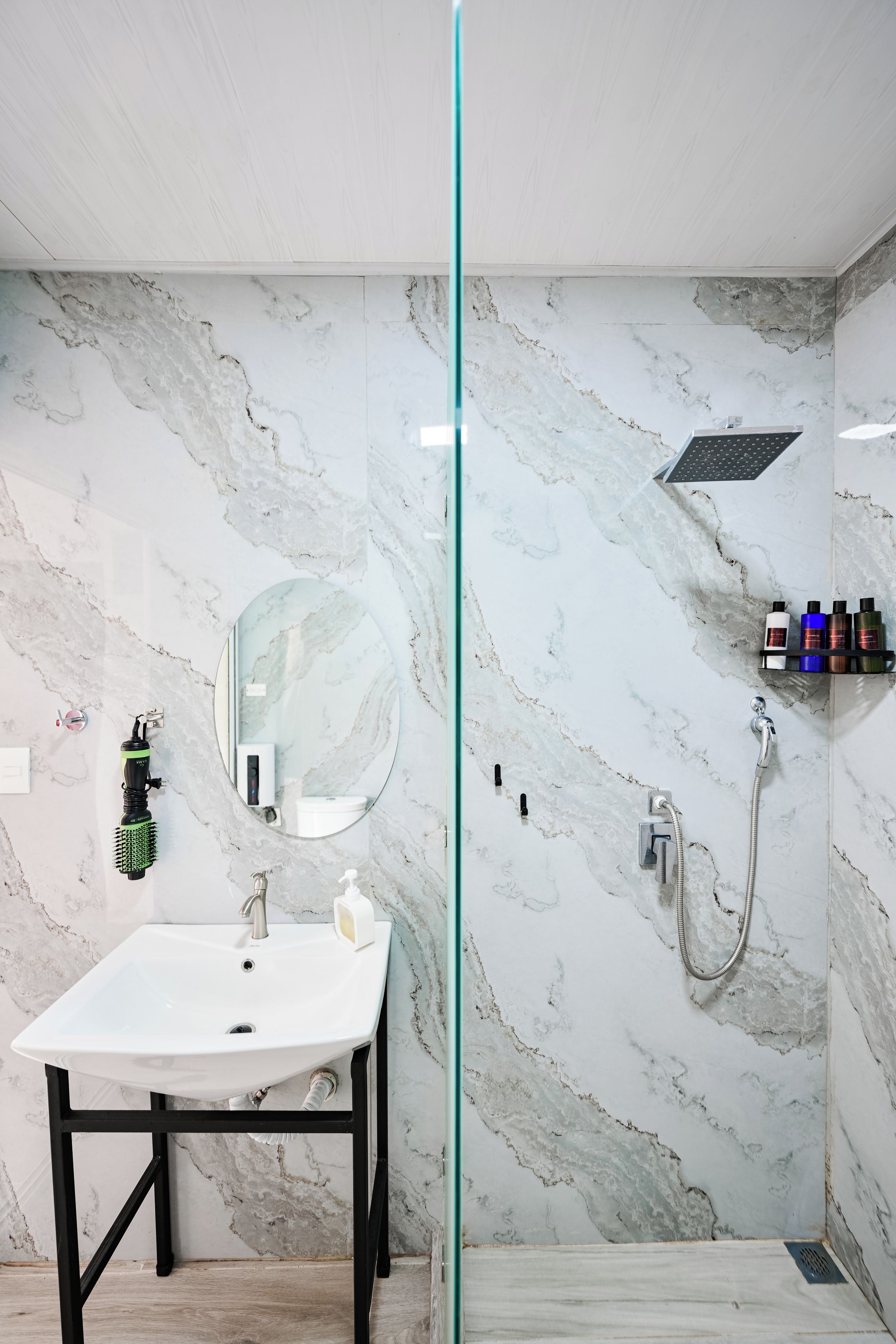 A bathroom with a marble wall, a white sink, a mirror, a shower, and a shelf with toiletries.