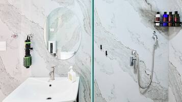 A bathroom with a marble wall, a white sink, a mirror, a shower, and a shelf with toiletries.