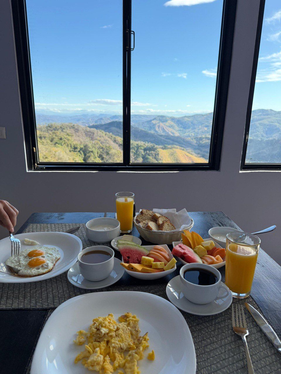 A breakfast table with eggs, toast, fruit, coffee, and juice, overlooking a mountainous landscape.