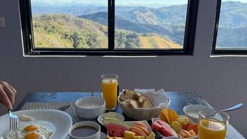 A breakfast table with eggs, toast, fruit, coffee, and juice, overlooking a mountainous landscape.