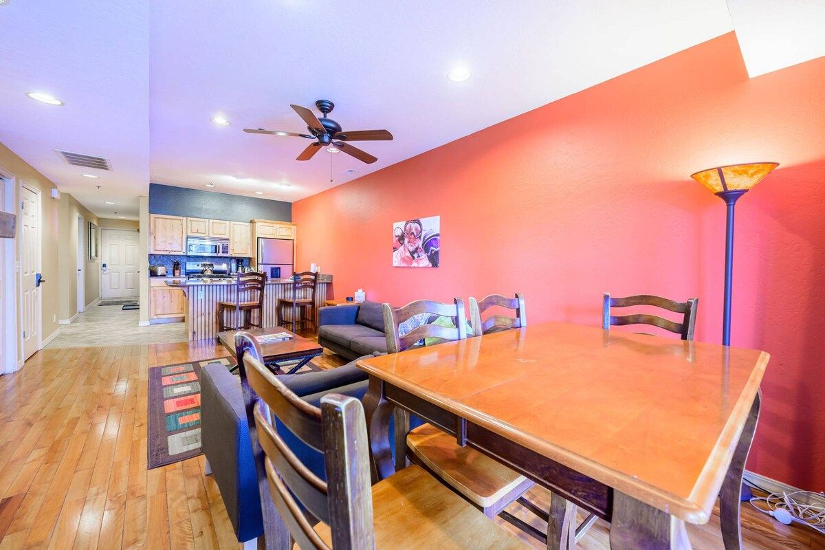 A dining area with a wooden table and chairs, a ceiling fan, and a kitchen in the background.