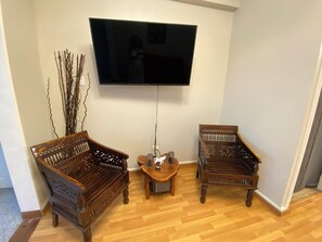 Un salon avec un téléviseur à écran plat fixé au mur, deux chaises en bois, une petite table sur laquelle est posé un vase, et une branche décorative.
