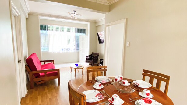 Un coin repas avec une table en bois dressée pour quatre personnes, entourée de chaises. On aperçoit une chaise rouge au fond, une fenêtre avec des stores et un luminaire au plafond.