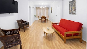 Een woonkamer met een rode bank, houten stoelen, een salontafel en een flatscreen televisie aan de muur.