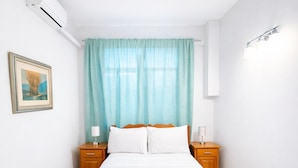 Een net opgemaakt bed met witte lakens en handdoeken, een houten hoofdbord, twee nachtkastjes met lampen, een raam met dunne gordijnen, en een ingelijste foto aan de muur.