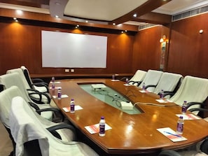 Une salle de conférence avec une grande table en bois, plusieurs chaises, un écran de projection et des bouteilles d’eau posées sur la table.
