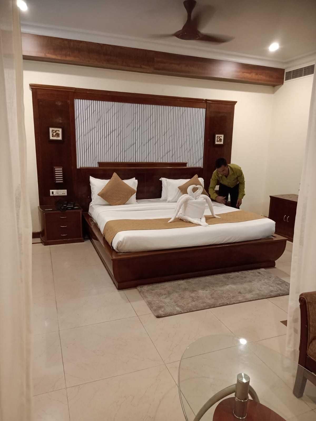 A hotel room with a large bed, wooden headboard, a nightstand, a bedside table, a ceiling fan, and a window with blinds.