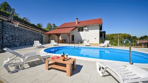 A swimming pool with lounge chairs, a wooden table with a bottle and glass, and a house with a red roof.