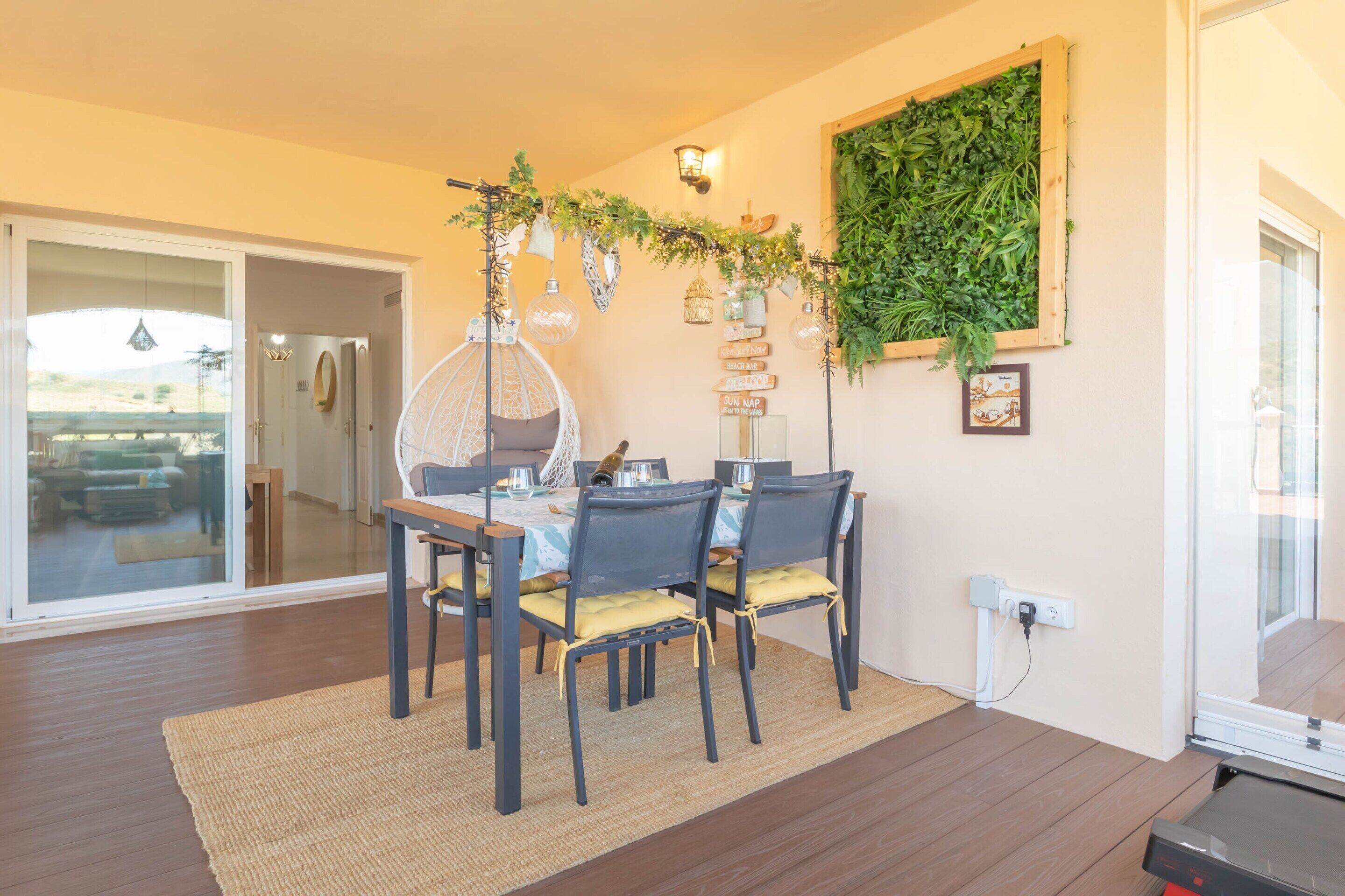 A dining area with a table set for four, surrounded by chairs with yellow cushions. A decorative garland hangs above the table, and a wall-mounted plant adds greenery to the space.