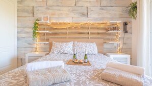 A bedroom with a wooden headboard, two bedside tables, a bed with patterned bedding, two pillows, a bedside table with a tray of champagne and a cupcake, and a hanging plant. - Mijas Golf Boutique Apartment - Golf & Leisure (Mijas)