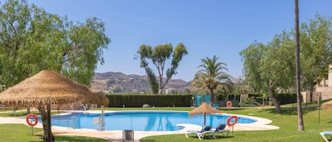 A swimming pool with lounge chairs and thatched umbrellas, surrounded by greenery and trees.