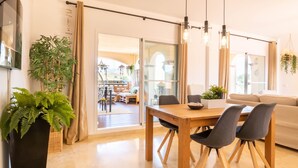 A modern living room with a wooden dining table, grey chairs, and a beige sofa. There are potted plants and a large window providing natural light. - Mijas Golf Boutique Apartment - Golf & Leisure (Mijas)