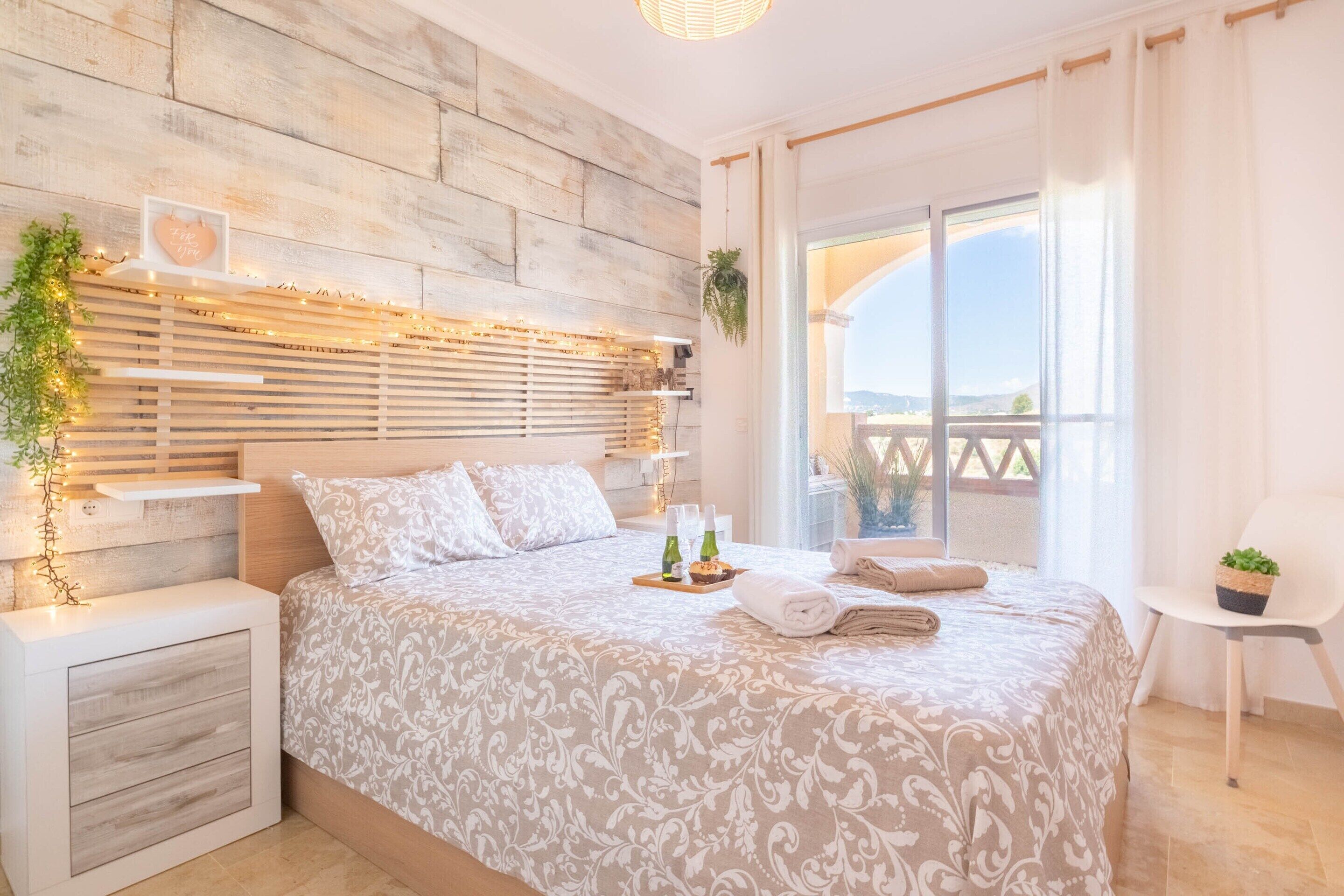A bedroom with a wooden headboard, a bed with patterned bedding, a bedside table with a plant, and a view of the outdoors through a large window.