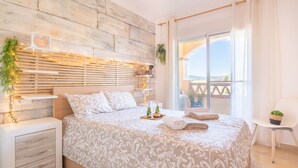 A bedroom with a wooden headboard, a bed with patterned bedding, a bedside table with a plant, and a view of the outdoors through a large window. - Mijas Golf Boutique Apartment - Golf & Leisure (Mijas)
