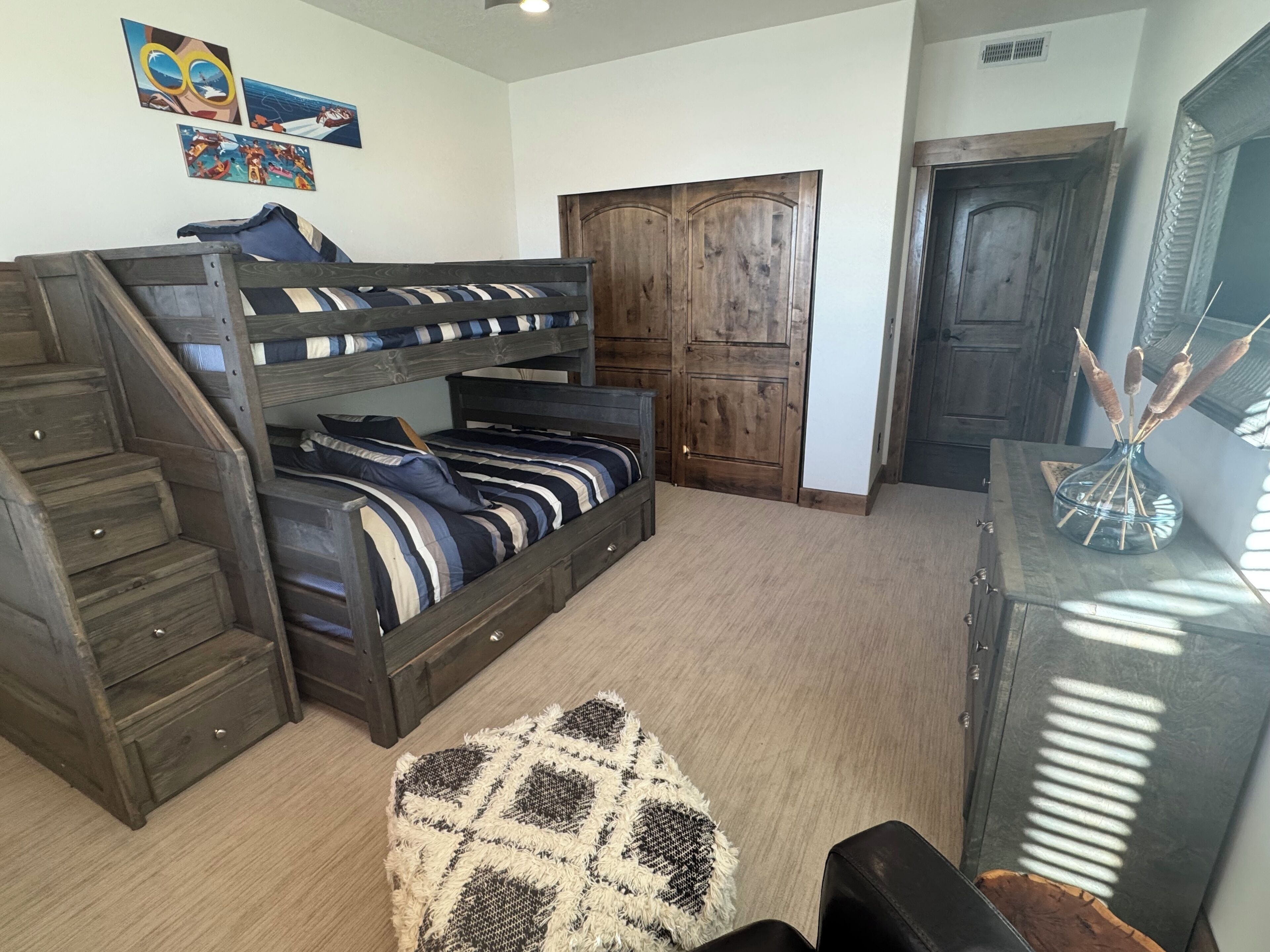 A bunk bed with a wooden ladder and drawers, a wardrobe, a desk with a vase, and a patterned rug.