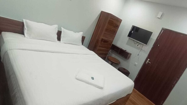 A hotel room with a large bed, a TV mounted on the wall, a wooden wardrobe, and a small table with a chair.