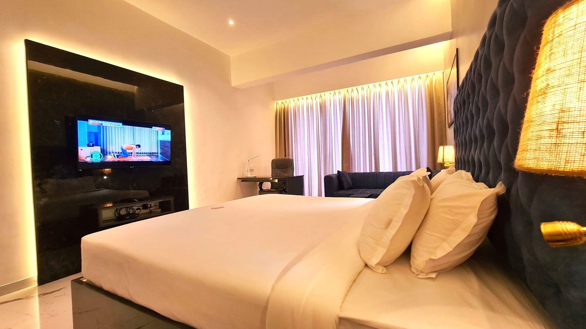 A modern hotel room with a large bed, a flat-screen TV, a desk with a chair, and a window with curtains.