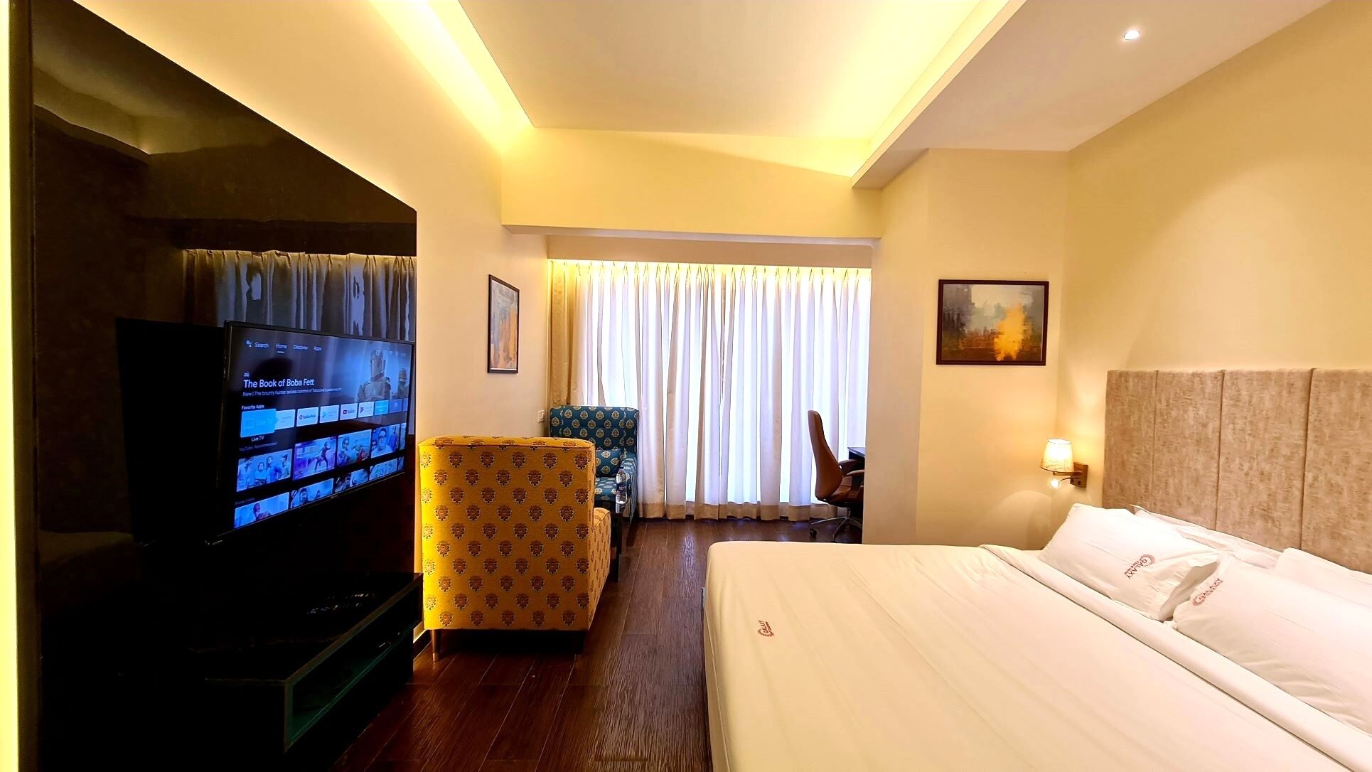 A hotel room with a bed, a television, a patterned armchair, a desk with a chair, and a painting on the wall.