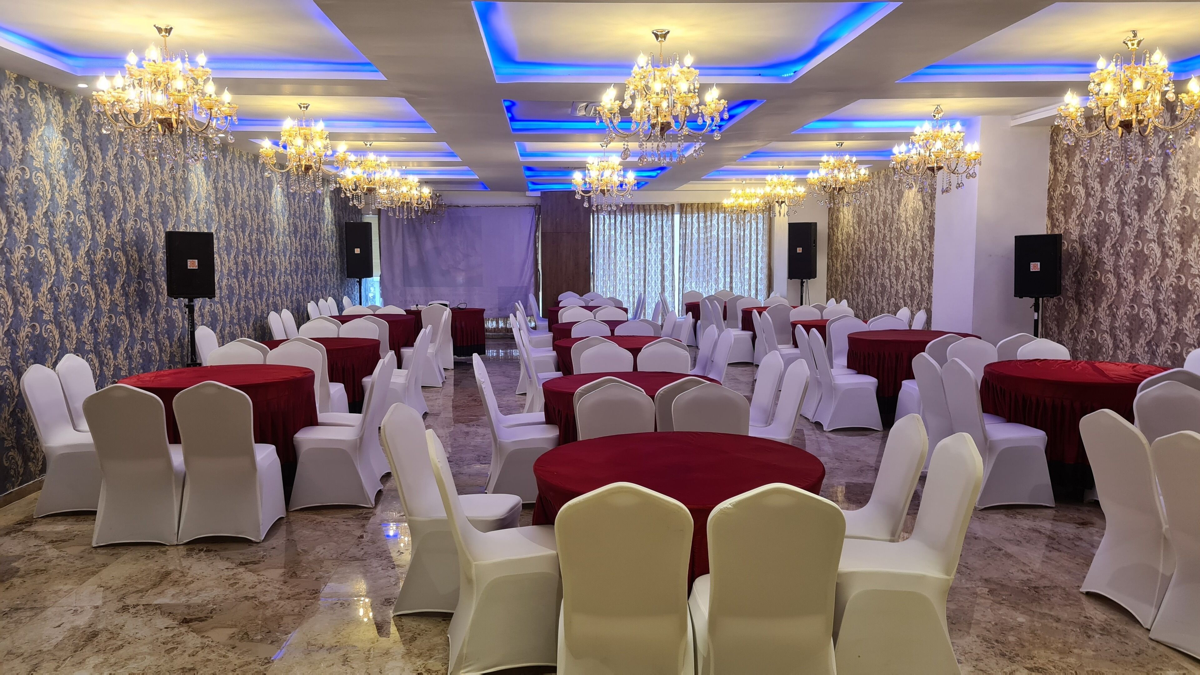 A banquet hall with round tables covered in red tablecloths, white chairs, and chandeliers.