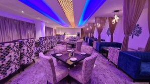 Un restaurante con zona de comedor central que muestra una mesa negra con platos y cubiertos blancos, rodeada de sillas tapizadas en color morado. Hay sofás azules y una pared divisoria con estampado.