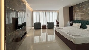 Habitación de hotel moderna con una cama grande, un televisor de pantalla plana montado en la pared, un rincón de descanso con dos sillones y un ventanal con cortinas.