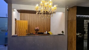 A hotel reception area with a wooden panel wall, a chandelier, and a reception desk with a fan and flowers. - Galaxy Club & Hotel (Bijapur)