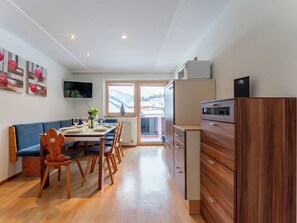 Uma cozinha moderna com armários de madeira, uma área de refeições com uma mesa posta para quatro pessoas e uma grande janela com vista para montanhas cobertas de neve.