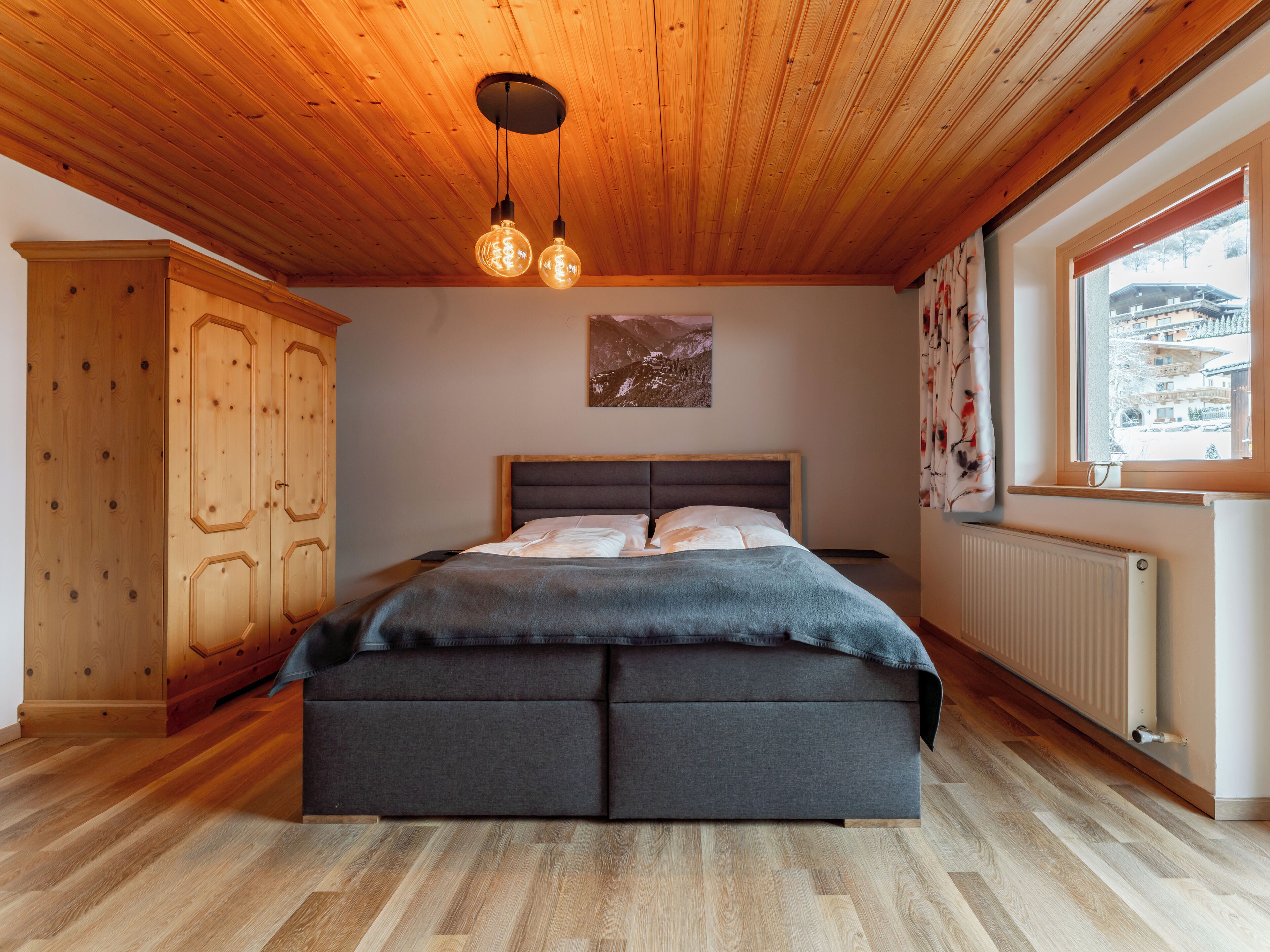 Een slaapkamer met een houten plafond, een bed met een grijze hoofdeinde en een raam met uitzicht op een besneeerd landschap.