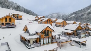 A cluster of wooden cabins with balconies, set on a snowy slope with mountains in the background.