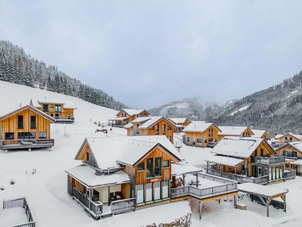 A cluster of wooden cabins with balconies, set on a snowy slope with mountains in the background.
