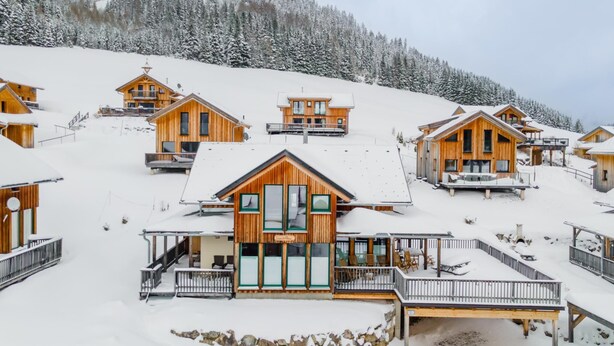 A snow-covered lodge with wooden cabins, a central building with large windows, and a stone retaining wall.