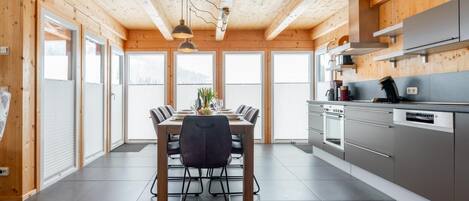 Una cocina moderna con vigas de madera, un comedor con mesa y sillas, y ventanas amplias.