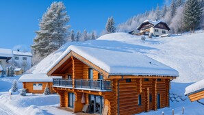 A log cabin with a balcony, surrounded by snow-covered trees and a clear blue sky.