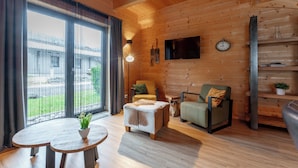Ein gemütliches Holzhaus-Interieur mit einer großen Schiebetür aus Glas, einem Sofa, einem Sessel, einem kleinen Tisch mit Pflanze und einem an der Wand befestigten Fernseher.